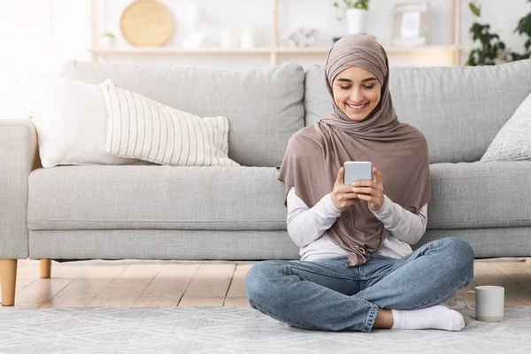 Modernes arabisches Mädchen im Hidschab checkt E-Mails auf dem Smartphone, sitzt auf dem Boden — Stockfoto