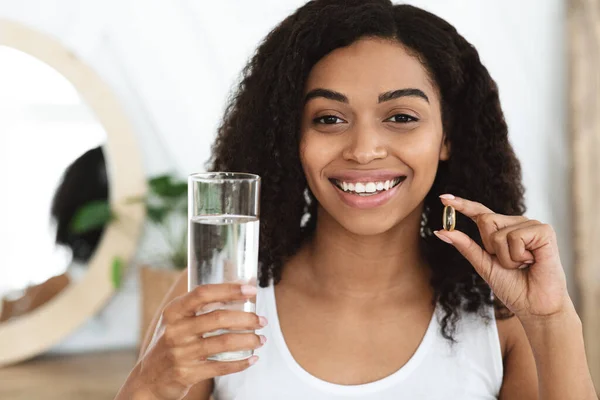 Vitaminas y complementos alimenticios. Sonriente mujer negra sosteniendo Omega-3 cápsula y agua —  Fotos de Stock