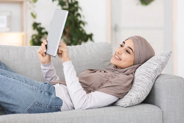 Sonriendo chica árabe en hijab usando tableta digital mientras se relaja en el sofá en casa, navegar por Internet o leer el libro electrónico — Foto de Stock