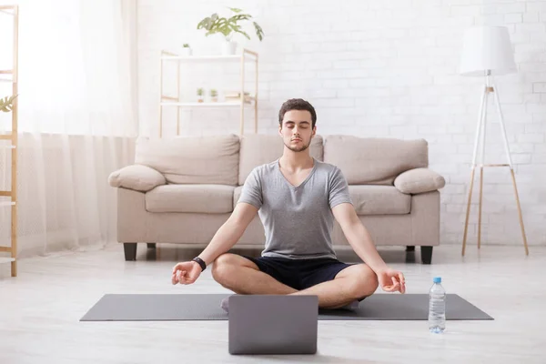 Medytacja i joga. Facet z zamkniętymi oczami siedzi w pozycji lotosu na macie, z laptopem — Zdjęcie stockowe