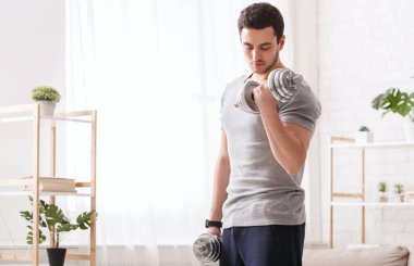 Spor salonu evde. Fitness izleyicisi olan adam dambıl tutuyor.