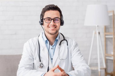 Evden çevrimiçi blog. Kulaklıklı gülümseyen doktor oturma odasının içindeki masada oturuyor.