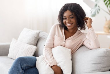 Yastıkla koltukta oturan, kameraya poz veren mutlu siyah kadın.