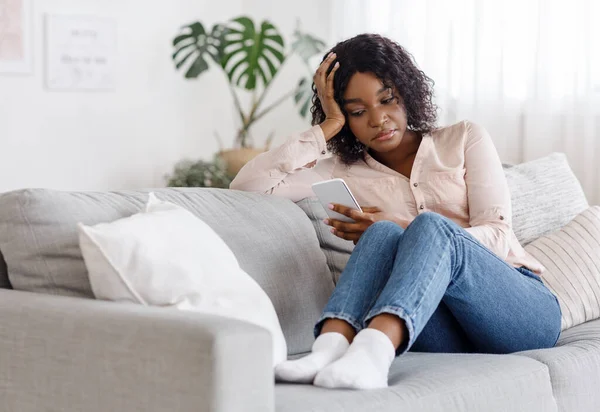Menina americana africana perturbada usando smartphone enquanto sentado no sofá em casa — Fotografia de Stock