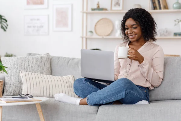 Концепция досуга. Молодая женщина отдыхает дома на диване, пьет кофе и использует ноутбук компьютер, копировальное пространство — стоковое фото