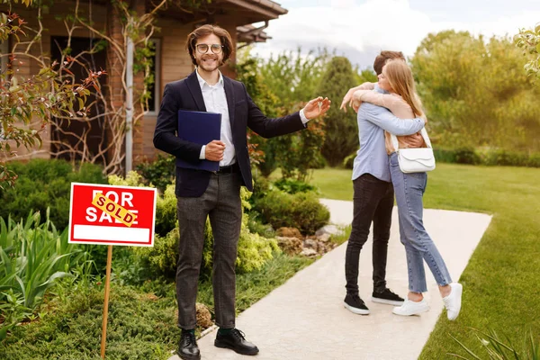 Smiling property manager and embracing millennial couple near beautiful house outdoors