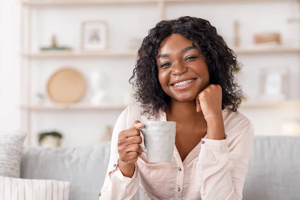 Vrolijke Afrikaanse vrouw ontspannen op de bank thuis met een kopje koffie — Stockfoto