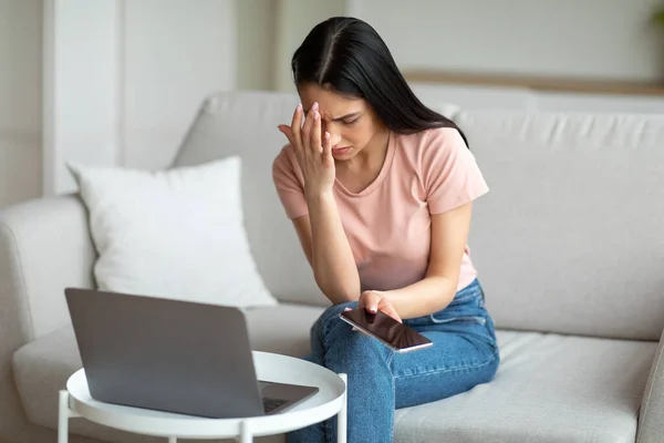 Mulher frustrada no laptop segurando telefone sentado no sofá interior — Fotografia de Stock