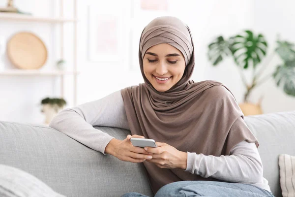 Réseaux sociaux. Sourire arabique fille dans la messagerie hijab sur smartphone à la maison — Photo