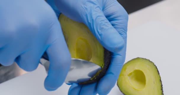Шеф-повар в перчатках снимает половину авокадо ложкой — стоковое видео