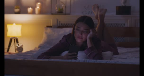 Unatkozó fiatal nő hálózatépítés mobiltelefon, feküdt az ágyon — Stock videók