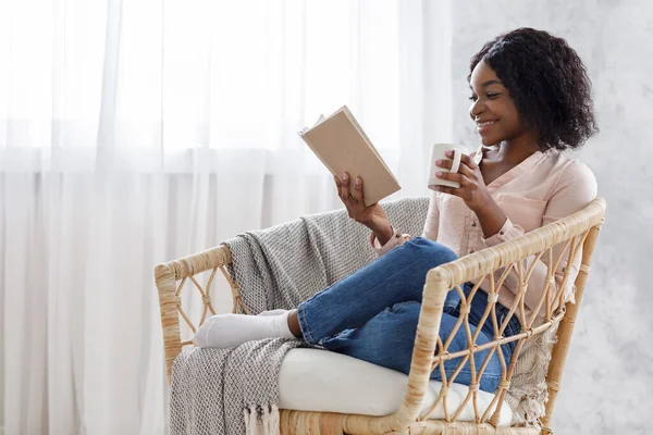 Pasatiempo de fin de semana. Joven mujer negra relajante en sillón con libro y café — Foto de Stock