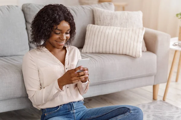 Online-Kommunikation. Glückliches schwarzes Mädchen beim SMS-Schreiben auf dem Handy, während es auf dem Boden sitzt — Stockfoto