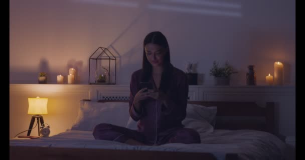 Веселая девушка разговаривает по телефону, сидит на кровати в пижаме — стоковое видео