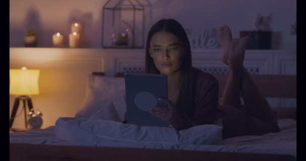 Serieuze vrouw die nieuws leest op digitale tablet, liggend op bed — Stockvideo