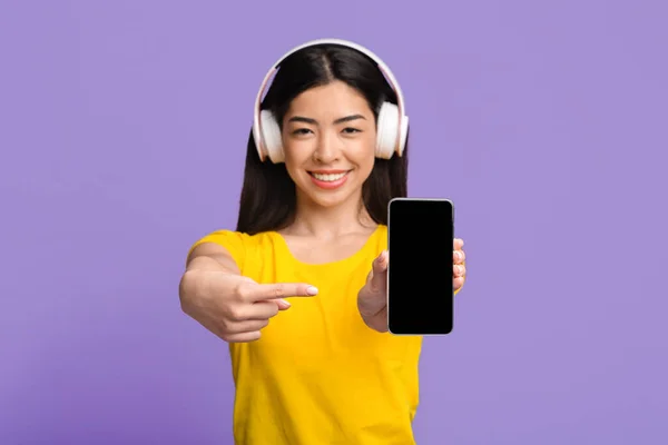 Service de streaming de musique. Souriant asiatique femme avec casque pointant sur smartphone noir — Photo