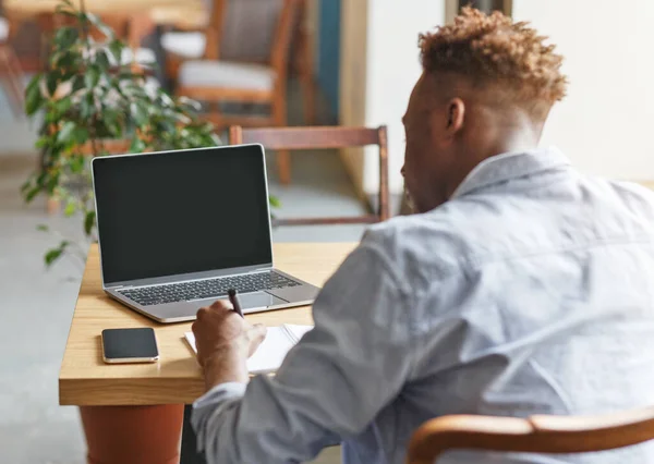Μαύρος τύπος γράφει κάτι στο σημειωματάριο κοντά στο laptop με άδεια οθόνη στο καφέ, mockup για το σχεδιασμό — Φωτογραφία Αρχείου