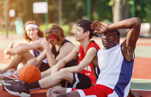 Exhausto jugador de baloncesto negro descansando con su diverso equipo en la cancha deportiva fuera — Foto de Stock