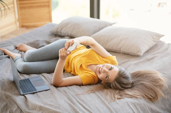 Jovencita alegre con ordenador portátil y teléfono móvil red de navegación en la cama en casa — Foto de Stock