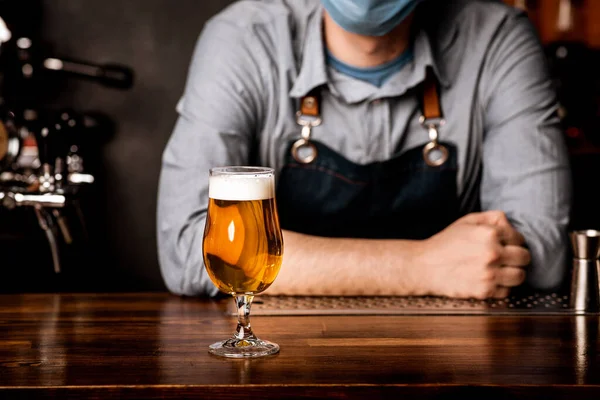 Bartender em máscara protetora se inclina no balcão de bar em que fica o vidro de cerveja leve com espuma — Fotografia de Stock