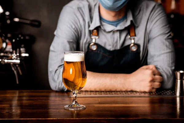 Cantinero en máscara protectora se apoya en el mostrador de la barra en la que se encuentra el vidrio de cerveza ligera con espuma — Foto de Stock