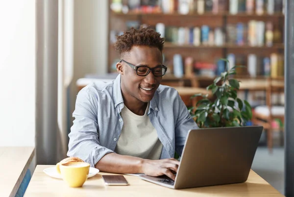 Alegre estudiante afroamericano haciendo su tarea en línea en la cafetería urbana — Foto de Stock