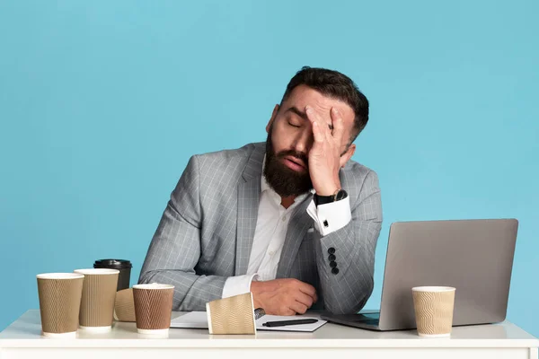A fazer horas extraordinárias. Empresário exausto sentado na mesa com laptop e copos de café vazios contra fundo azul — Fotografia de Stock