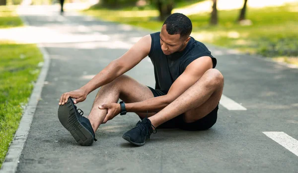 Sportverletzungen. Afroamerikanischer Läufer sitzt auf Joggingstrecke und spürt Schmerzen im Knöchel — Stockfoto