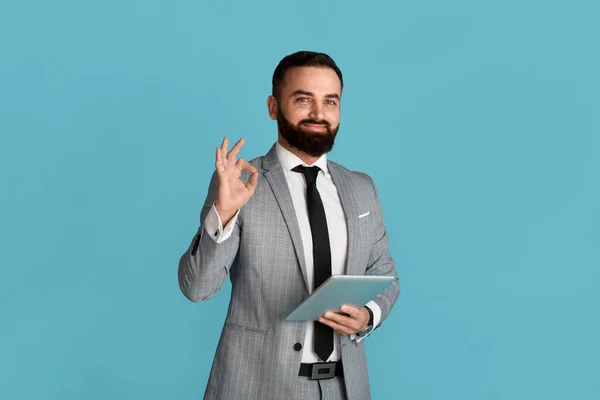 Retrato de hombre de negocios guapo con tableta que muestra el gesto OK sobre fondo azul —  Fotos de Stock