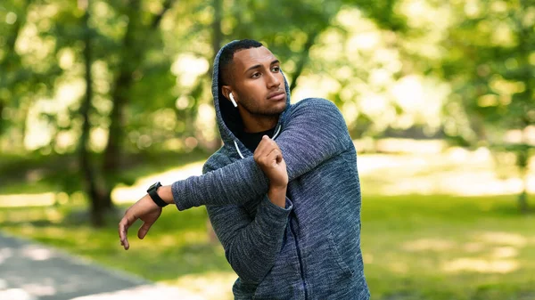 Αθλητικός Αφροαμερικανός με κουκούλα που τεντώνει το χέρι του και ακούει μουσική μετά την προπόνηση στο καλοκαιρινό πάρκο — Φωτογραφία Αρχείου