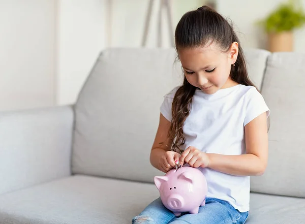 Menina colocando moeda em Piggybank sentado no sofá em casa — Fotografia de Stock