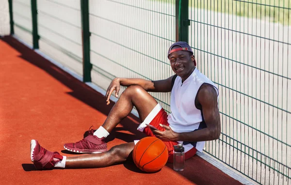 Esportista preto bonito com bola e telefone celular tendo descanso na quadra de basquete ao ar livre — Fotografia de Stock