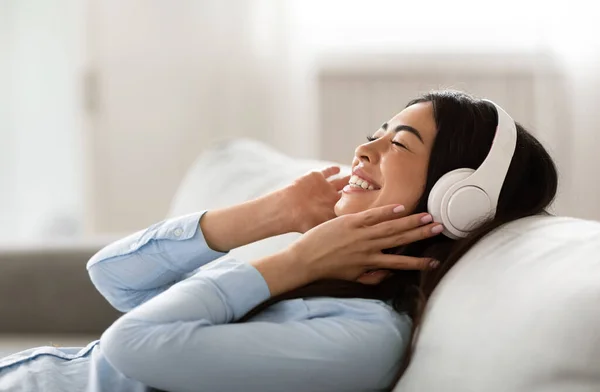 Ocio favorito. Relajada chica africana escuchando música en auriculares inalámbricos con los ojos cerrados, acostado en el sofá en casa, Vista lateral — Foto de Stock