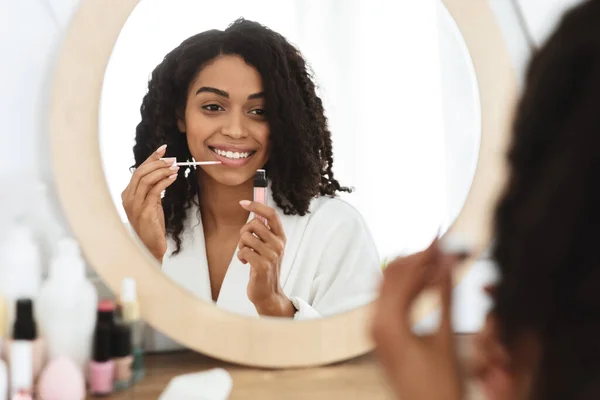 Rutina de maquillaje. Sonriente chica afroamericana aplicando brillo de labios cerca del espejo —  Fotos de Stock