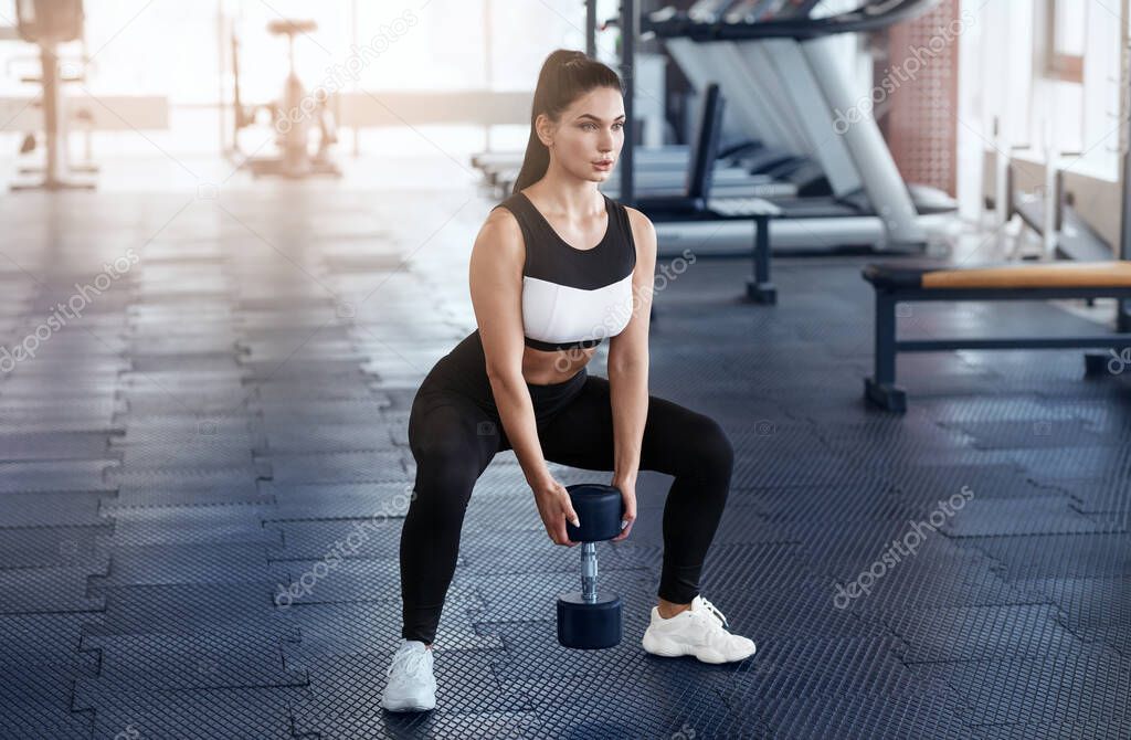 Strong young woman doing squats with dumbbell in sports club