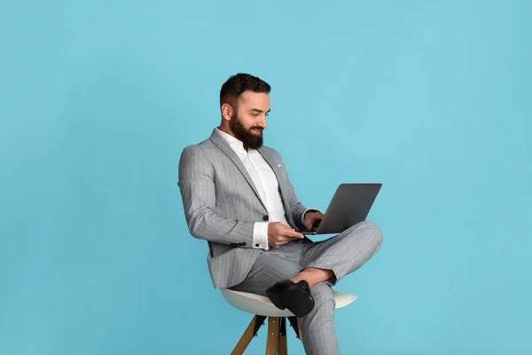 Aantrekkelijke zakenman met behulp van laptop computer terwijl zitten op stoel tegen een blauwe achtergrond — Stockfoto