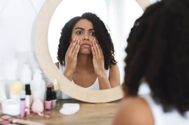 Anti-Wrinkle Skincare. Upset Black Woman Touching Her Face, Looking In Mirror clipart
