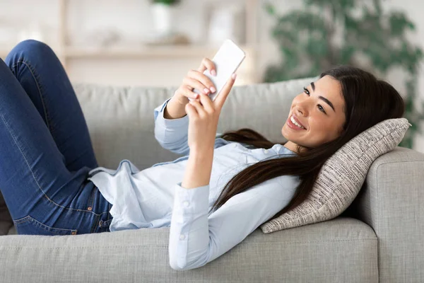 Giovane donna asiatica messaggistica su smartphone a casa, sdraiata su un comodo divano — Foto Stock