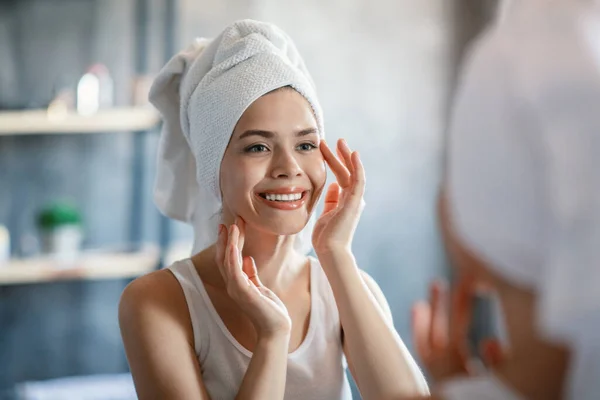 Feliz mujer milenaria con toalla en la cabeza y piel radiante frente al espejo en el baño — Foto de Stock