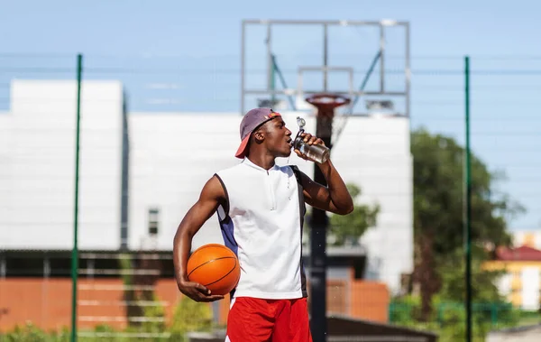 Pemain basket muda berkulit hitam minum air untuk menyegarkan diri setelah berlatih di lapangan terbuka — Stok Foto