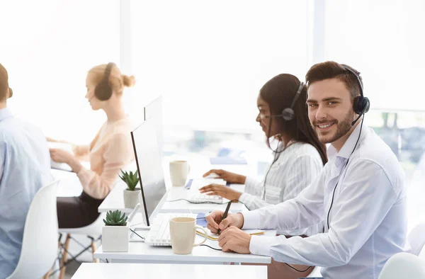 Atractivo operador de centro de llamadas sonriendo a la cámara y sus compañeros de trabajo hablando con los clientes en la oficina, espacio de copia —  Fotos de Stock