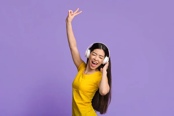 Joyeuse fille asiatique dans les écouteurs sans fil danser et chanter sur fond violet — Photo