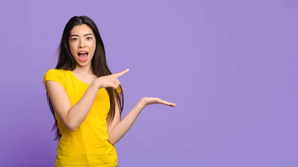 Emocionado chica asiática señalando en Open Palm, Demostrando algo sobre fondo púrpura — Foto de Stock