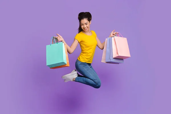 Веселая азиатская шопоголичка прыгает с большим количеством пакетов покупок на фиолетовом фоне — стоковое фото