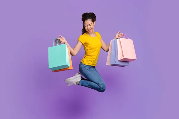 Vrolijke aziatische shopaholic meisje springen met veel shopping tassen over paarse achtergrond — Stockfoto