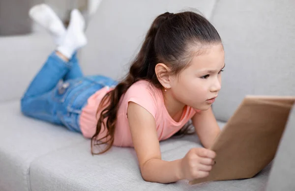 Pequeña chica asiática leyendo libro acostado en sofá en casa —  Fotos de Stock