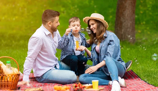 Lindo chico con sus padres divirtiéndose en el picnic, soplando burbujas de jabón en el parque — Foto de Stock