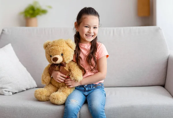 Asiática niña abrazando oso de peluche sentado en sofá interior — Foto de Stock