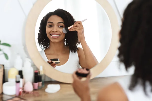 Hermosa mujer negra aplicando rubor en la cara, usando herramienta de cepillo, poniendo maquillaje — Foto de Stock