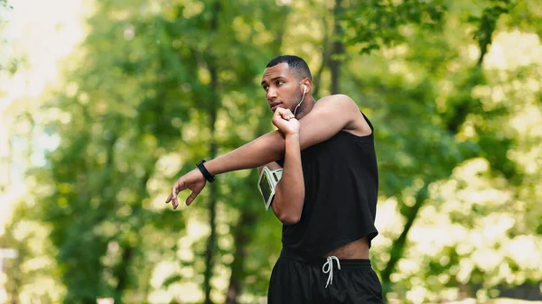 Cool black guy with earphones stretching his arm and listening to music on his morning workout at park, empty space — Stock Photo, Image